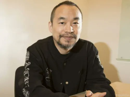 A man with a short beard and shaved head, wearing a black long-sleeved shirt with white graphics on the sleeves, sits and smiles slightly at the camera. The post context suggests this is "Machi Big Brother," the trader known as the "King of Liquidations" on Hyperliquid, whose dramatic trading losses and comeback are being discussed.