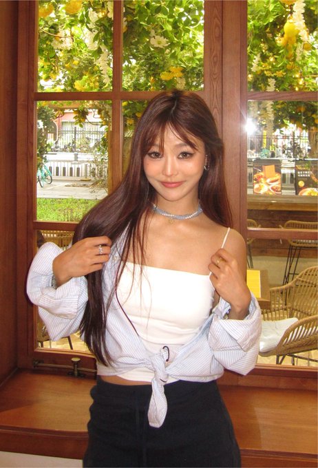 A woman with long dark hair posing indoors in front of a window, wearing a white top tied at the waist with a striped shirt and a black skirt, accessorized with a sparkly choker.