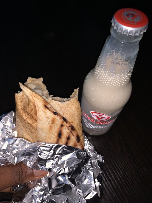A hand holds a foil-wrapped grilled wrap next to a chilled glass bottle of Yanaamilk with a red cap, the contrast of warm street food and cold milk suggesting a casual meal break amid the count down to the NomismaNetwork launch in 7 days.