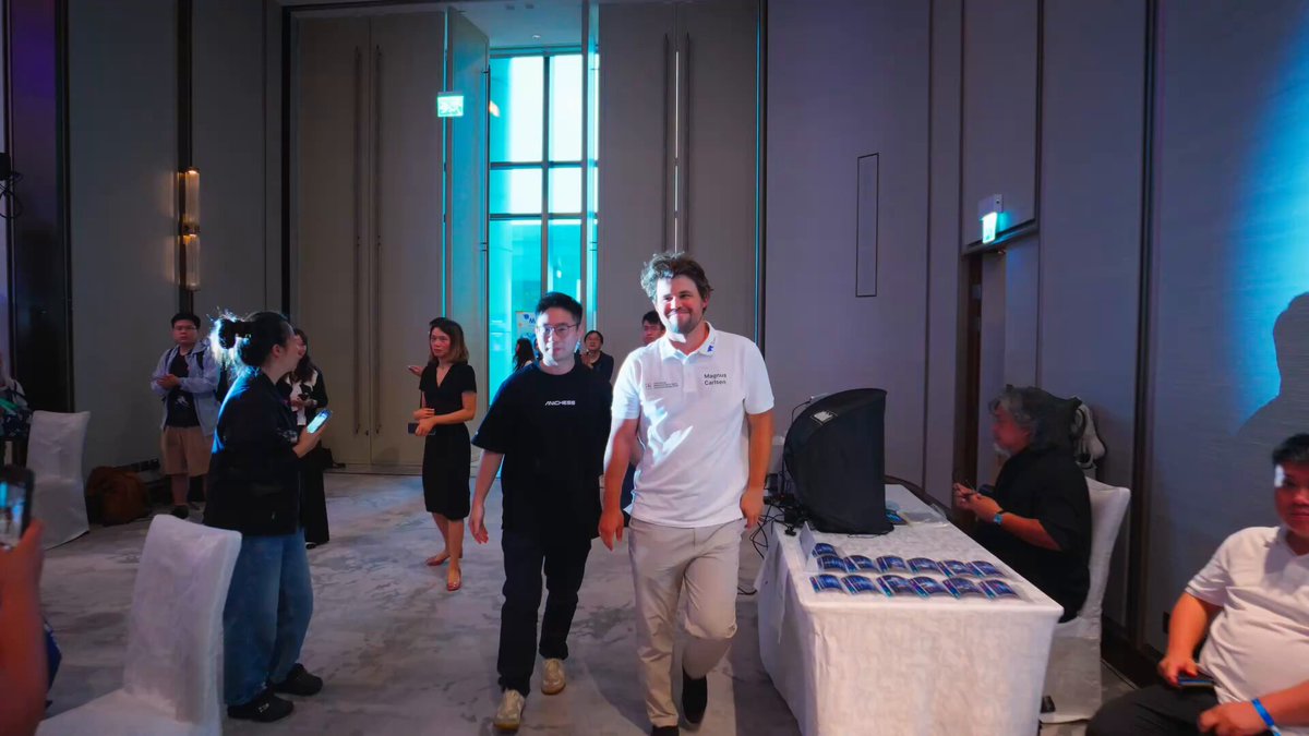 The image shows a group of people walking down a hallway towards a table with a computer monitor at its center. The people appear to be engaged in conversation or preparing for an event. There are several chairs placed around the room, indicating that this is likely a gathering space or meeting area. A person wearing a white shirt can also be seen in the image.