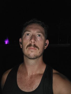 A close-up photo of a man with a mustache wearing a black tank top in a dark setting, with a purple light visible in the background.