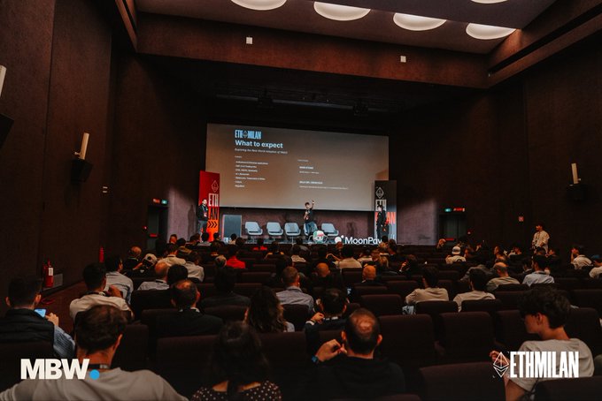 A presentation taking place in a movie theater-style auditorium with a large screen displaying "CTM Milan What to expect," watched by a seated audience.