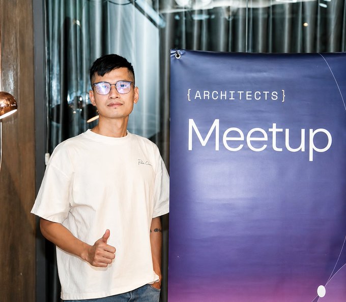 An Asian man wearing glasses and a white t-shirt gives a thumbs-up next to a purple banner that reads '{ARCHITECTS} Meetup' at what appears to be a developer community event.