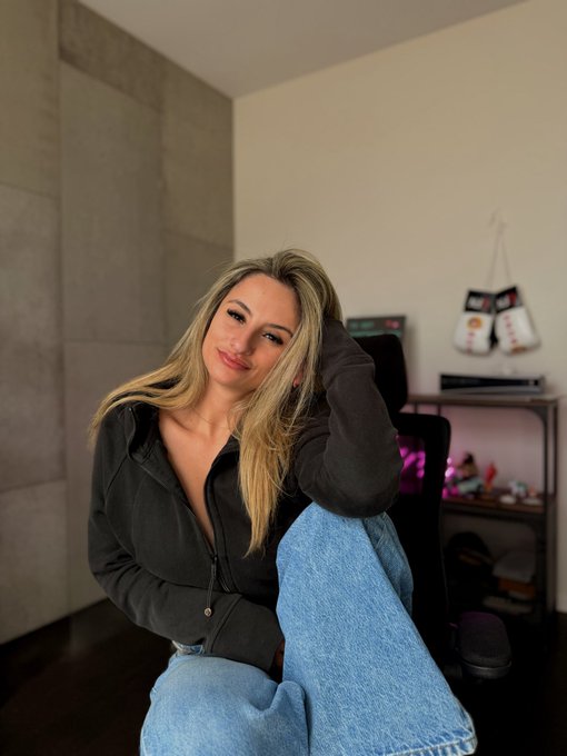 A blonde woman in a dark zip-up hoodie and blue jeans poses indoors, with boxing gloves hanging on the wall and a neon sign visible in the background.