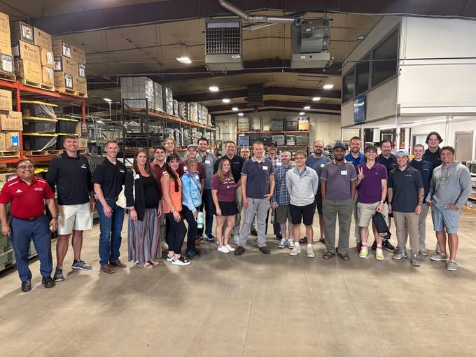A large group of approximately 25 employees stands posing for a photo inside an industrial warehouse or factory, surrounded by tall racks filled with cardboard boxes and equipment.