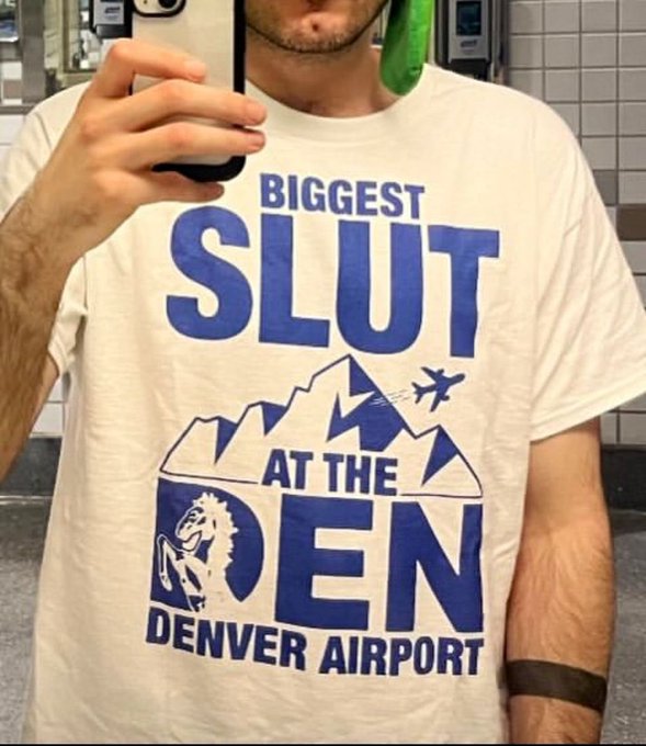 A person takes a mirror selfie wearing a white t-shirt that reads 'BIGGEST SLUT AT THE DEN — DENVER AIRPORT' with a mountain graphic and airplane.
