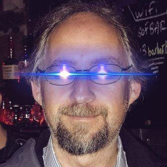 A close-up photograph of a man with glasses, a goatee, and long hair, with bright blue light reflections visible on his lenses and a chalkboard menu in the background.