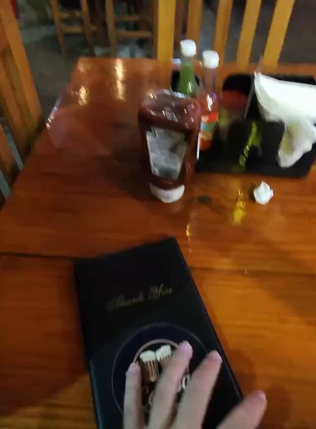 The image shows a person's hand resting on top of a black notebook, which appears to be a menu or a receipt from a restaurant. The background features a dining table with various condiments such as ketchup bottles and a small container of sauce. There are also chairs around the table, suggesting that it is likely a restaurant setting.