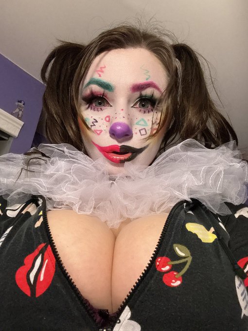 A person in clown makeup with colorful designs on their cheeks, a purple nose, and a black lips detail, wearing a white ruffled collar and a patterned zip-up top.
