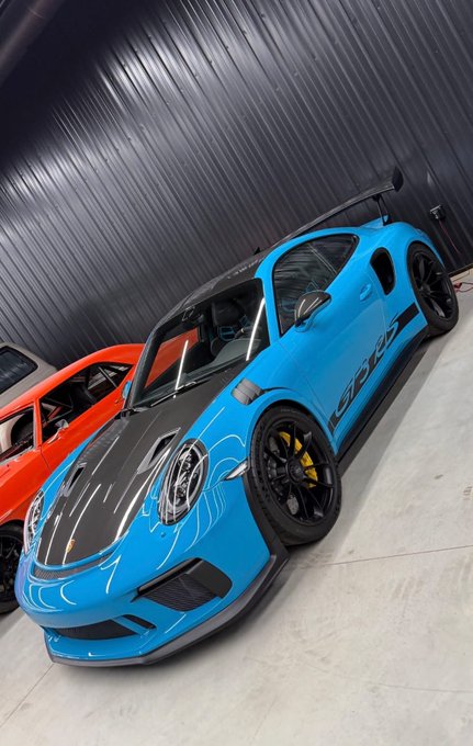 A bright blue Porsche 911 GT3 RS with black accents and a large rear wing is parked in a garage next to an orange car. The sports car features "GT3 RS" badging on its side, fitting the luxury automotive theme likely shared in the post thanking ANSEM.