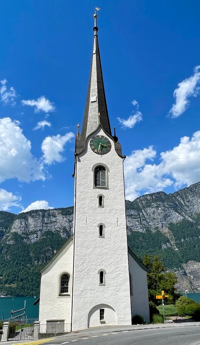 A tall white church with a pointed spire and clock tower, set against a backdrop of steep mountains and a lake under a blue, partly cloudy sky.