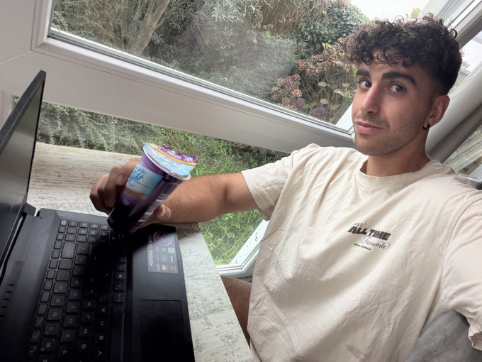 A young man in an "ALL TIME" t-shirt sits at his laptop near a window, holding up what appears to be a yogurt or dessert cup with a spoon, looking at the camera with a slight smirk—illustrating the casual, on-the-go multitasking of someone juggling ChatGPT queries and crypto market checks while grabbing a snack.
