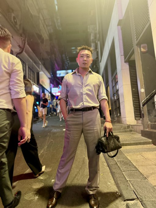 A young Asian man in a light-colored button-up shirt and gray trousers stands on a city street at night holding a black leather bag.