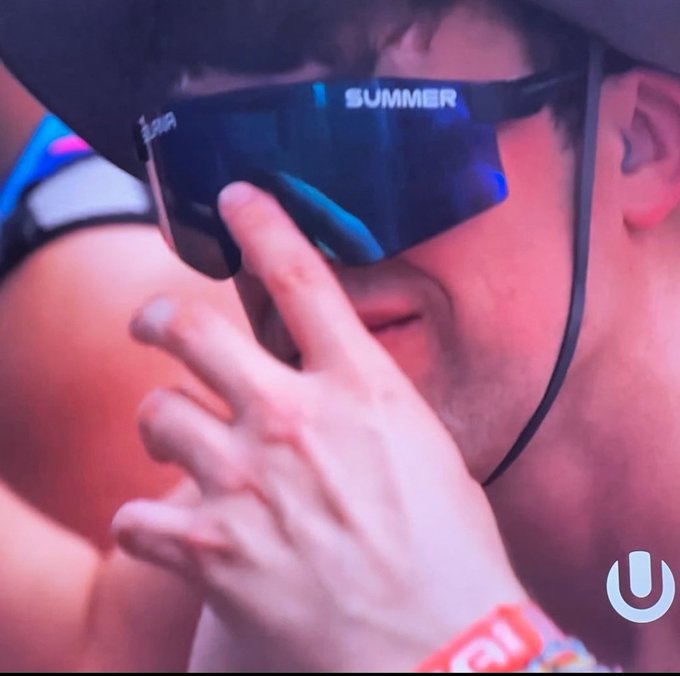 A close-up photograph of a person wearing reflective sunglasses with the word 'SUMMER' visible on the frame, with a finger raised near their face and a festival wristband visible on their wrist.