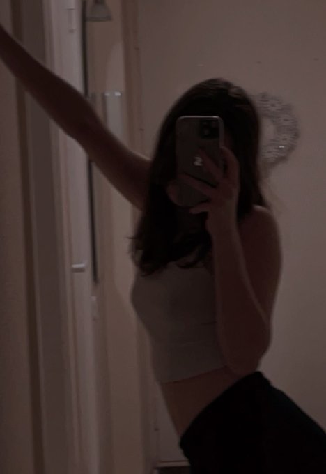A young woman takes a mirror selfie in dim lighting with her iPhone, wearing a tan crop top and dark pants, with one arm raised.