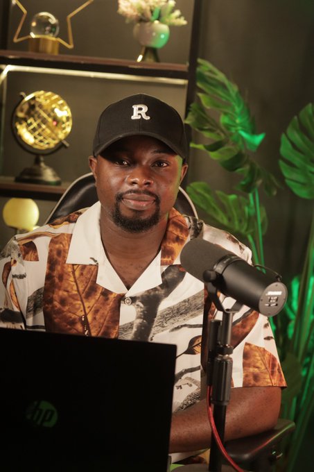 A man wearing a black cap with an 'R' logo and a patterned shirt sits in front of a microphone in a studio setting with plants and decorative lighting.