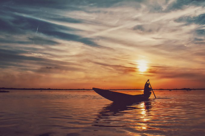 The image features a person rowing a boat out into the water at sunset, with the sun setting behind them. The sky above has beautiful orange hues that create a stunning backdrop for the scene. The person appears to be enjoying their time on the water as they navigate through the calm waters under the warm glow of the setting sun.