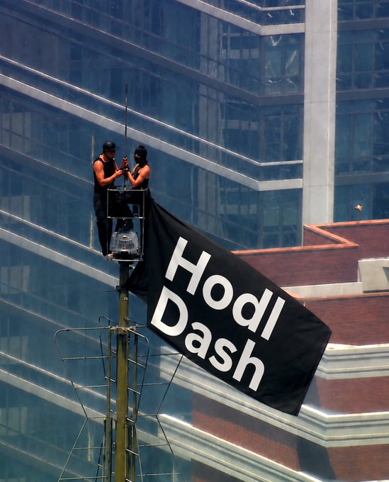 Two people on a tower platform near a glass office building hold a black banner with white text reading 'HODL Dash'.
