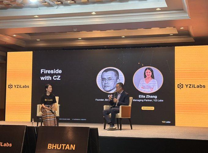 Two individuals sit on stage for a "Fireside with CZ" panel at a YZi Labs event in Bhutan, with a large screen displaying CZ (Founder, Binance) and Ella Zhang (Managing Partner, YZi Labs). The company logo "YZi Labs" is visible on both side panels, matching the post's mention of a discussion between CZ and Ella Zhang about blockchain, AI, and founder lessons.