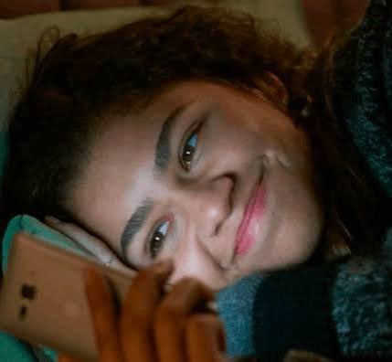 A close-up portrait shows a smiling young woman with thick eyebrows looking up while holding a smartphone, wearing a teal textured sweater.