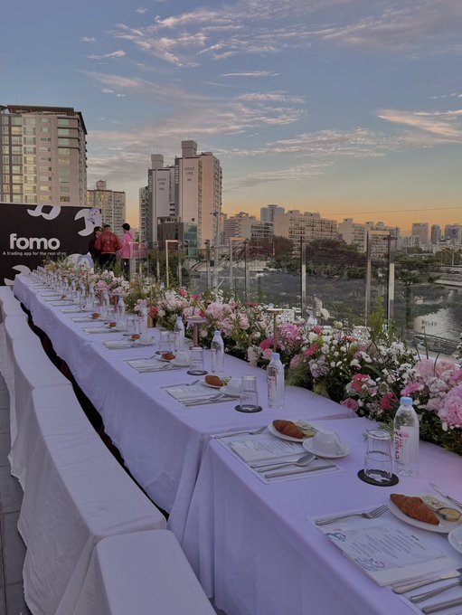 An elegant rooftop dinner setup with a long white-clothed table adorned with pink and white floral centerpieces, place settings, and a 'fomo' banner, overlooking a city skyline at sunset.
