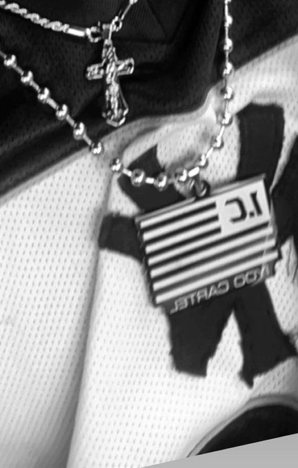 A black and white close-up photograph of silver chains, a cross pendant, and a 'Turbo Cartel' logo charm resting on a textured fabric surface.