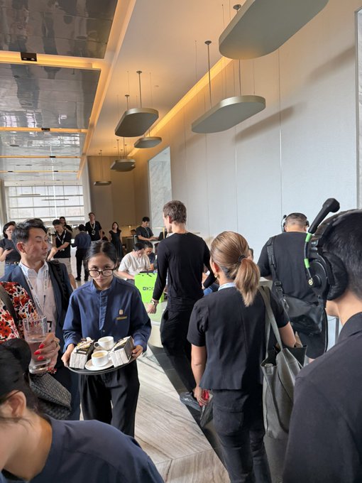 A server in a blue uniform carries a tray of coffee and pastries through a crowded, modern event space with staff and guests.