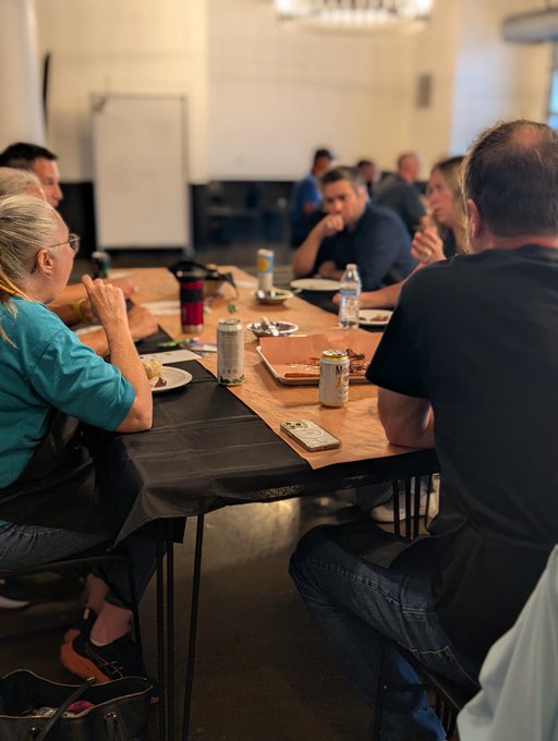 A group of people sit around a long wooden table eating food and drinking canned beverages in what appears to be a casual gathering or event.