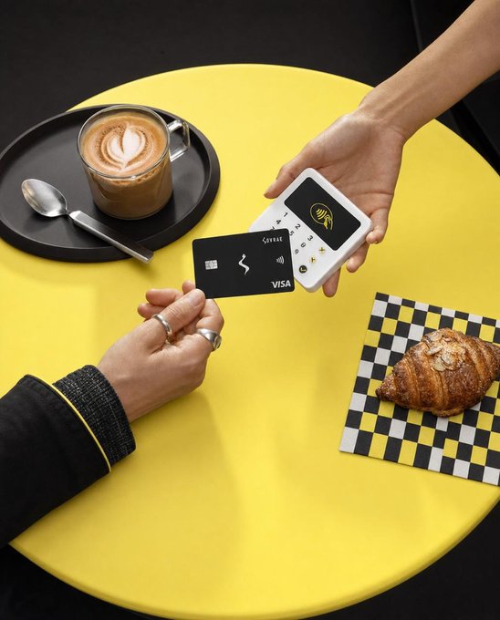 An overhead shot of a contactless payment transaction on a yellow table, showing a hand holding a card reader accepting a black Visa card, with a cappuccino, croissant, and checkered napkin nearby.