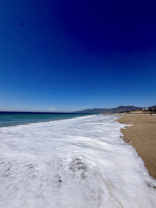 A scenic beach view with white foamy waves washing onto tan sand under a clear blue sky, with mountains visible in the distance.