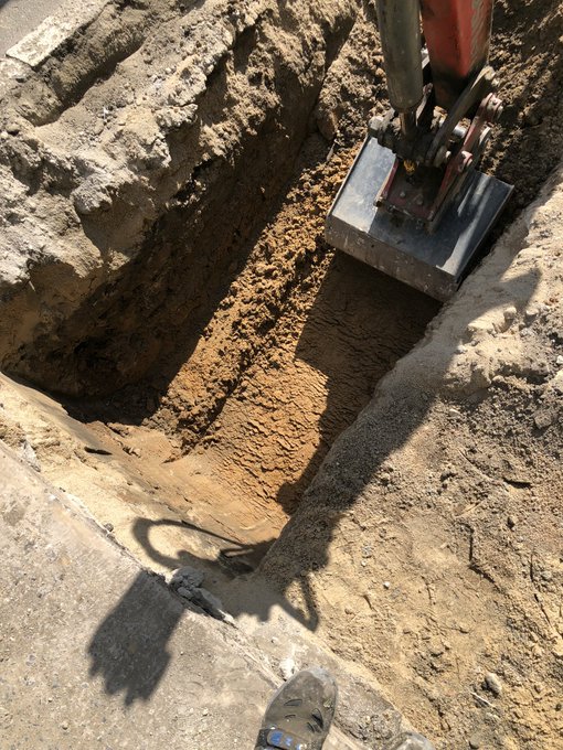 An excavation site showing an excavator bucket digging a trench in dirt, with a worker's boot and a cable visible at the bottom of the frame.