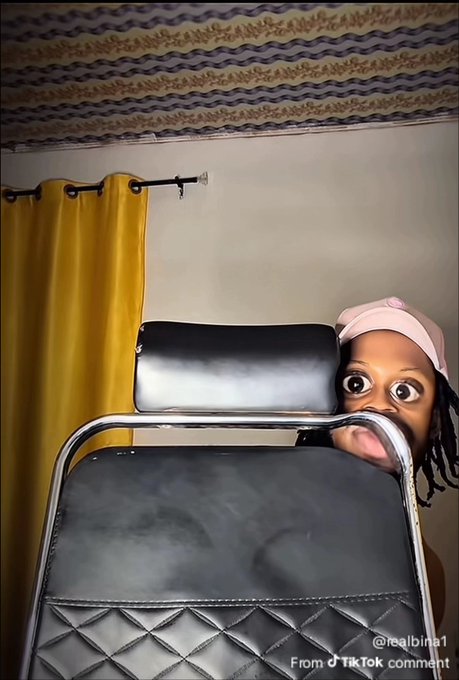 A person wearing a pink head covering peeks out playfully from behind a black padded chair in a room with yellow curtains and patterned ceiling fabric, with a TikTok watermark '@realbina1' reading 'From a TikTok comment'.