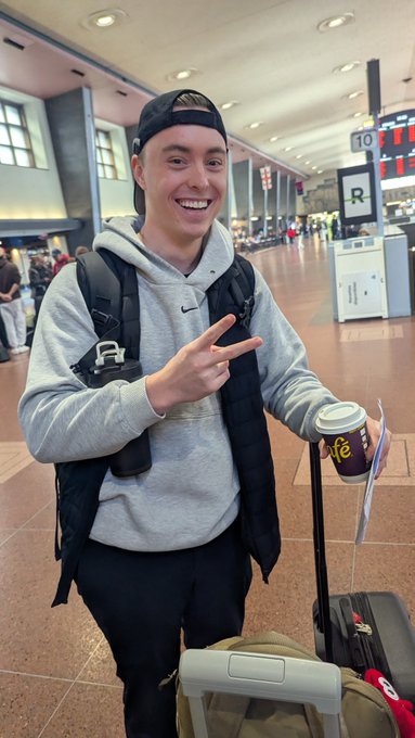 A smiling young man in a gray Nike hoodie and backwards cap flashes a peace sign while holding a "Life" coffee and his suitcase handle at a train station, with gate number "10" and an "R" sign visible in the background—perfectly capturing the casual excitement of spotting a friend before boarding.