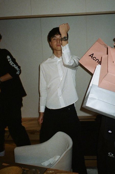A man in a white shirt and black trousers raises an arm indoors, with partially visible people and a large Acne Studios shopping bag around him.