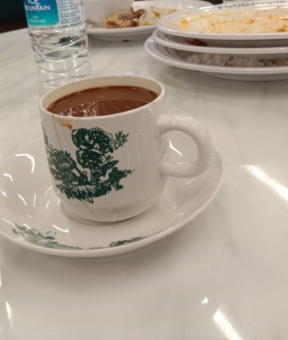 A vintage-style teacup filled with coffee or tea sits on a matching saucer on a marble table, with a bottle of Ice Mountain water and stacked plates of food in the background.
