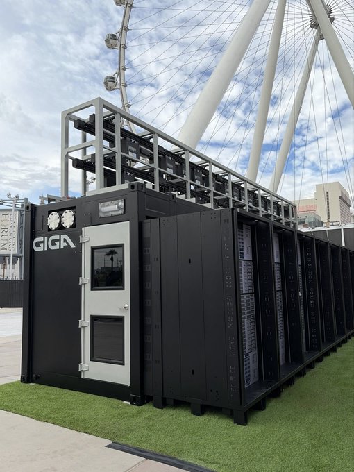 A black container-like data center structure branded with "GIGA" is displayed on artificial grass, featuring a door and exhaust fans on top, set against a large Ferris wheel.