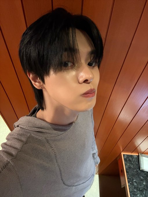 Close-up selfie of a young man with dark hair wearing a gray textured hoodie, taken in a room with wood paneled walls.