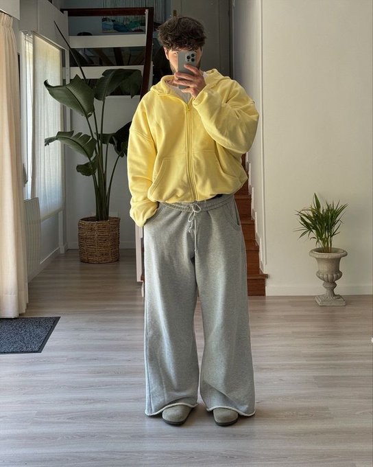 A person taking a mirror selfie wearing a yellow zip-up hoodie and oversized gray sweatpants in a bright room with houseplants.