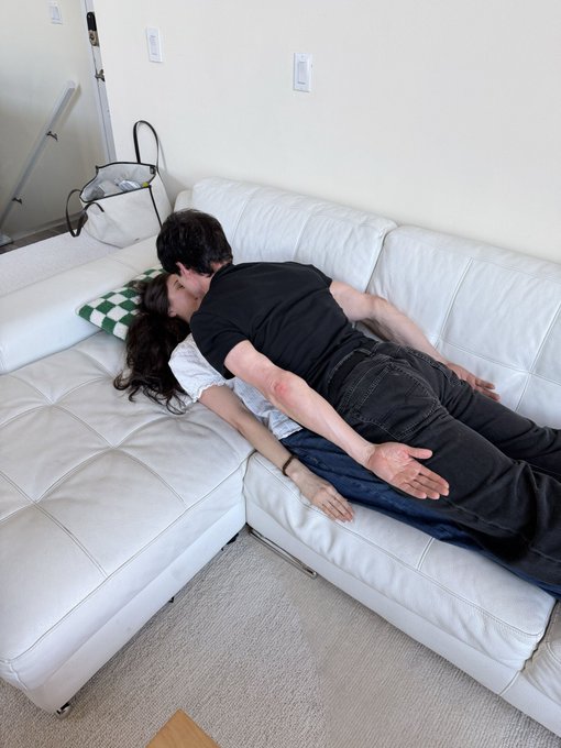 A couple relaxes on a white leather sectional couch in a living room, with one person lying across the other.