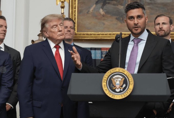 President Donald Trump stands beside a man speaking at a podium bearing the Presidential Seal in what appears to be the White House, with other officials and a large painting visible in the background.