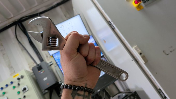 A tattooed hand grips a chrome adjustable wrench in an industrial setting with electrical control panels visible in the background.