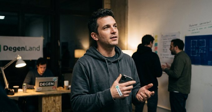 A man in a gray hoodie speaks at an event with a 'DegenLand' sign visible in the background, while others work at desks with laptops.