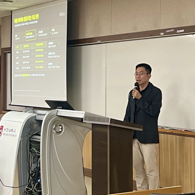 A man in glasses and dark clothing speaking into a microphone at a podium during a lecture, with a presentation slide displayed on a screen behind him showing Korean text about business trends.