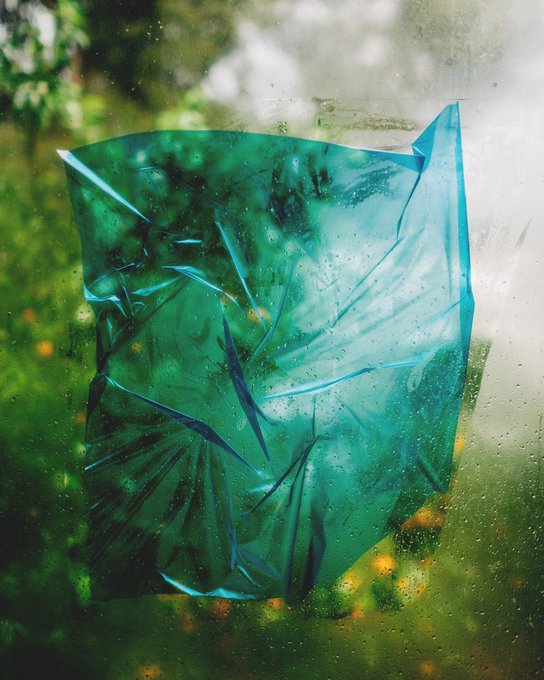 A crumpled piece of translucent teal plastic pressed against a rain-speckled window, with blurred green foliage and warm orange bokeh lights visible through the glass. The text "rainy day" and "{[ 5 / 8 ]} ~~ 8.88 xtz" indicates this is a digital artwork edition being celebrated by its creator for being collected.