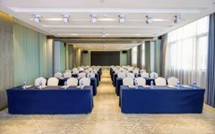 An empty conference or meeting room with rows of chairs covered in blue fabric arranged facing a front podium area, lit by natural light from large windows.