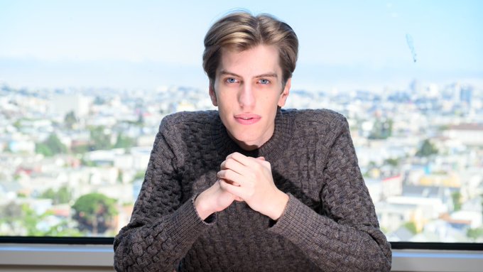 A young man in a brown cable-knit sweater sits with hands clasped, looking directly at the camera against a backdrop of a hazy cityscape, conveying a pensive, defeated expression that mirrors the post's narrative of financial ruin.