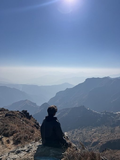 A person sits on a rocky outcrop, silhouetted against a vast mountain range under a bright, clear sky, taking in the expansive sunrise view. The image complements the post's peaceful, reflective tone, evoking a serene weekend moment in nature that contrasts with the digital world of web3.