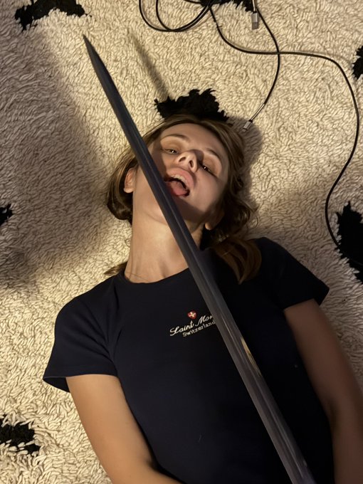 A young woman with blonde hair lies on a fluffy white and black rug, playfully holding a sword up to her open mouth as if pretending to swallow it, wearing a dark blue t-shirt with partially visible "Saint Mar" text. The image ties to the "$dasha" post with its edgy, dramatic pose suggesting a bold or risqué social media aesthetic.