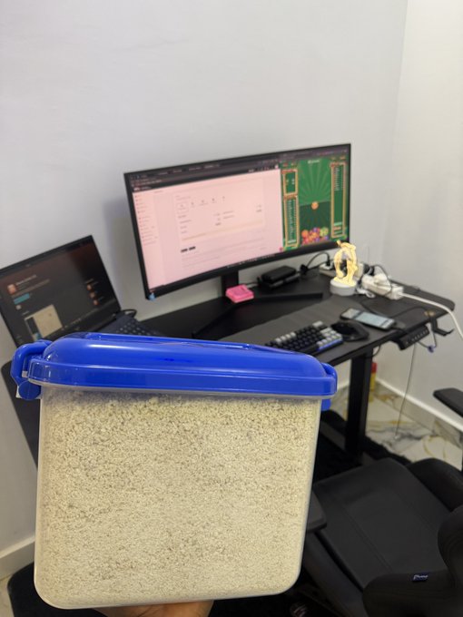 A hand holding up a large clear plastic container with a blue lid filled with what appears to be dried herb or seasoning, in front of a computer desk setup.