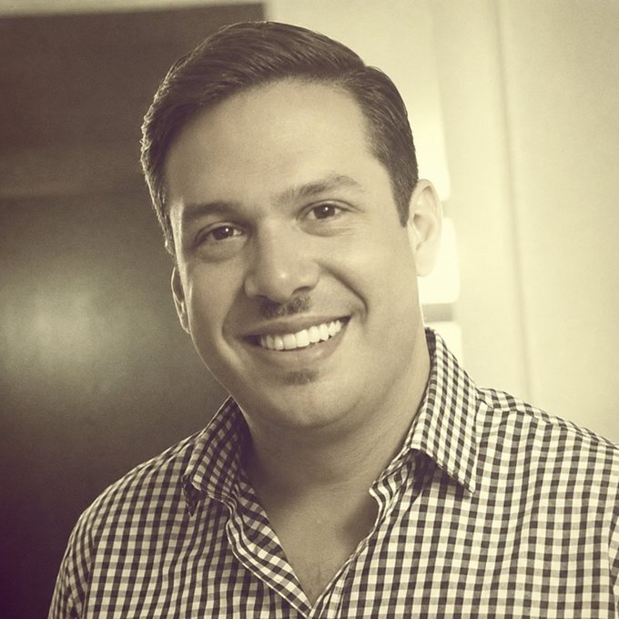 A man with a wide smile wears a checkered collared shirt, his warm and friendly expression matching Adolfo's introductory tone as he seeks name suggestions for his new Bitcoin hardware company.
